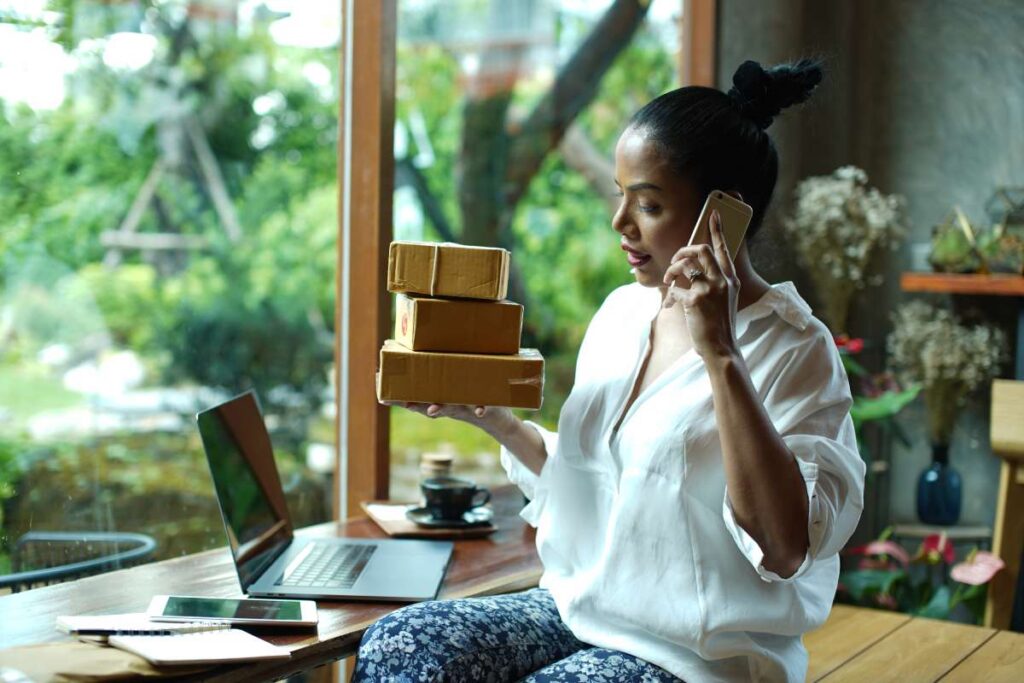 A young woman speaking on her phone while holding a stack of boxes.